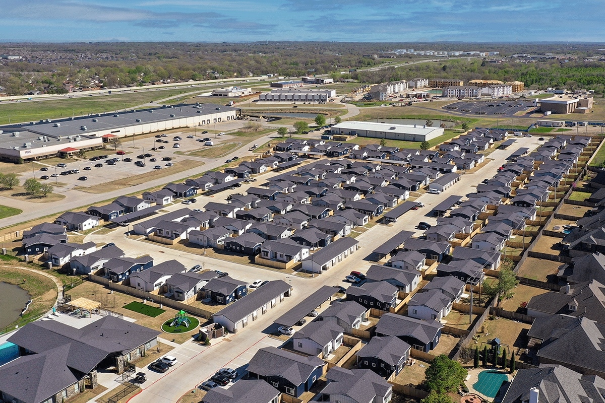Aerial view of a suburban neighborhood with rows of houses, streets, parking lots, nearby commercial buildings, and a pond in the lower left corner on a clear day.