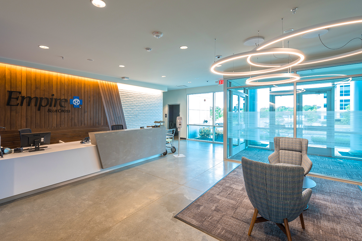 A modern office lobby with a reception desk, two armchairs, circular hanging lights, large glass entrance doors, and a sign reading Empire BlueCross on a wood accent wall.