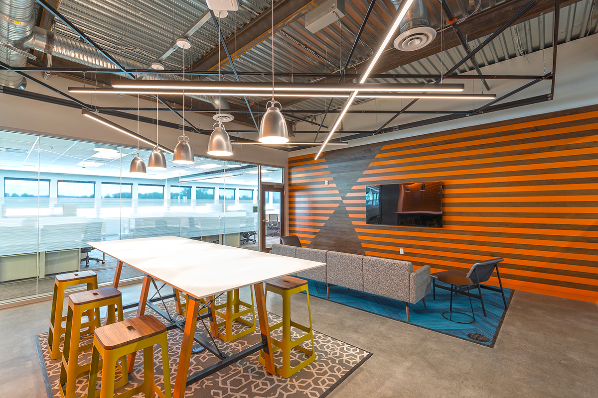 Modern office lounge with a striped orange and brown accent wall, TV, gray sofa, blue rug, high table with yellow stools, and sleek pendant lights under an exposed metal ceiling. Glass wall shows adjacent office space.