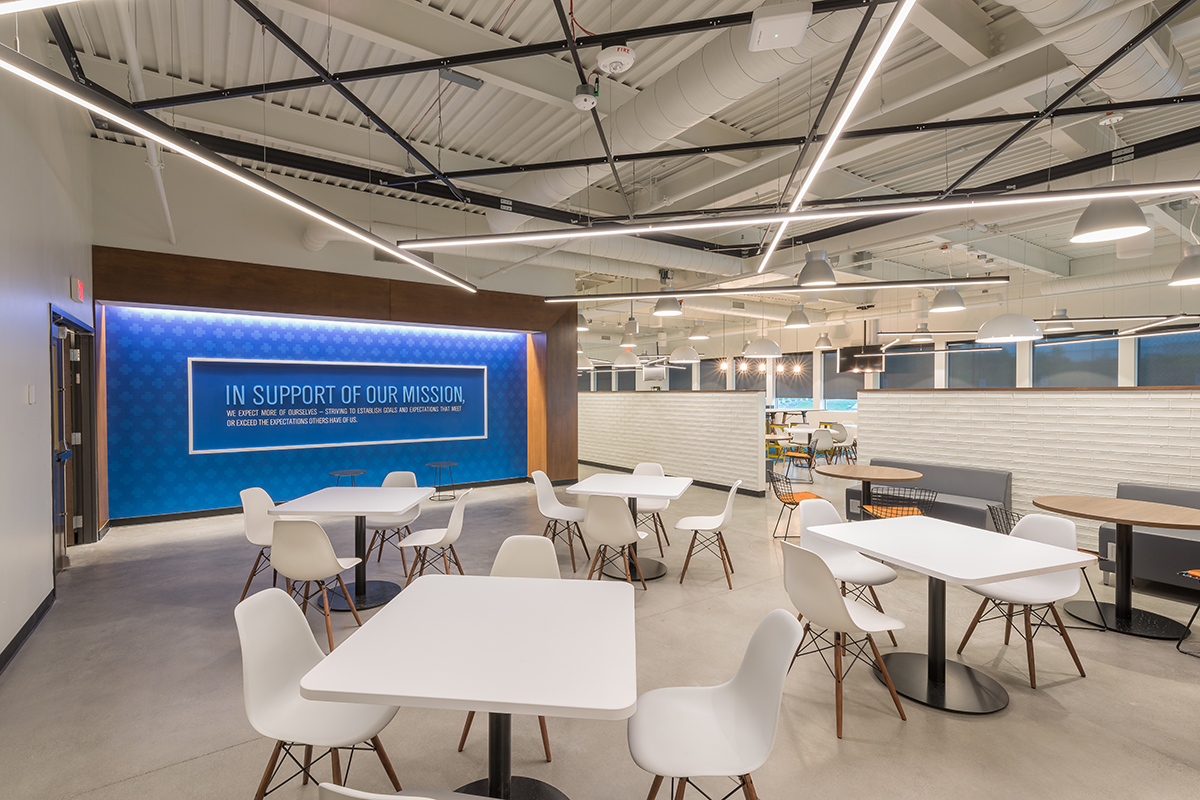 Modern office break room with white tables and chairs, overhead lighting, and a blue wall featuring the text In Support of Our Mission. Partition walls separate the seating area from the rest of the office.