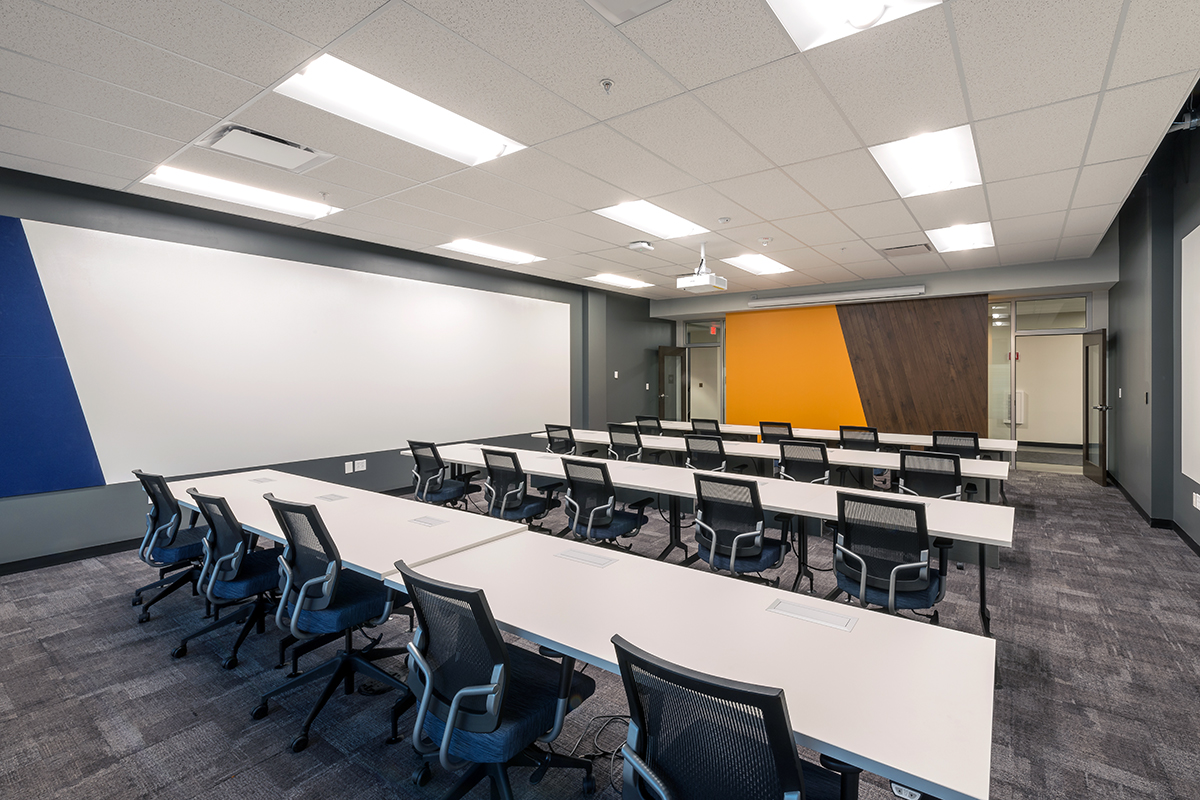 Modern classroom with rows of white desks, black office chairs, a large whiteboard, projector, and colorful accent walls in blue and orange. The room is empty and well-lit with ceiling lights.