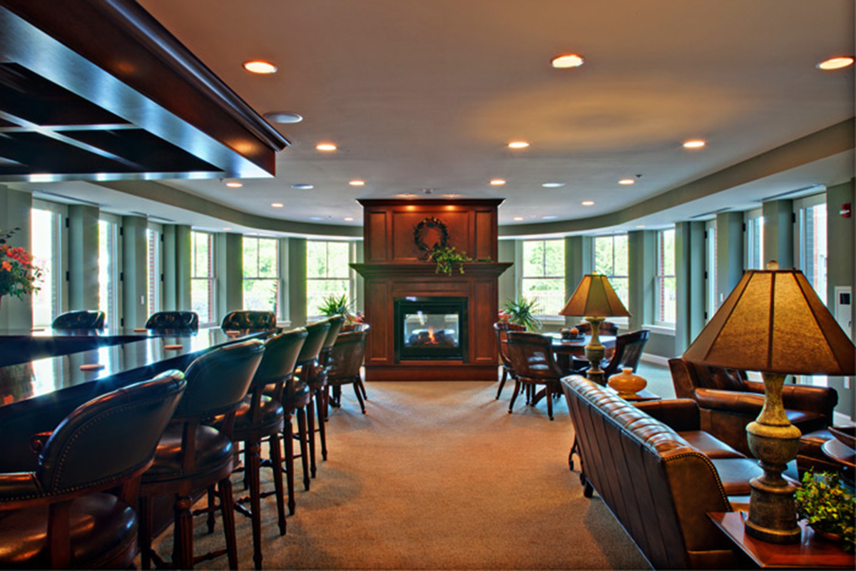 A spacious, upscale lounge with leather chairs, sofas, a fireplace, bar seating, and large windows letting in natural light. Warm lighting and wood accents create a cozy, inviting atmosphere.