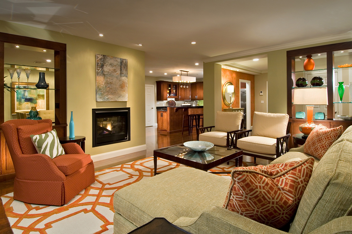 Modern living room with beige sofa, orange armchair, patterned rug, glass coffee table, and open shelves displaying vases. A fireplace and kitchen are visible in the background. Warm lighting creates a cozy atmosphere.