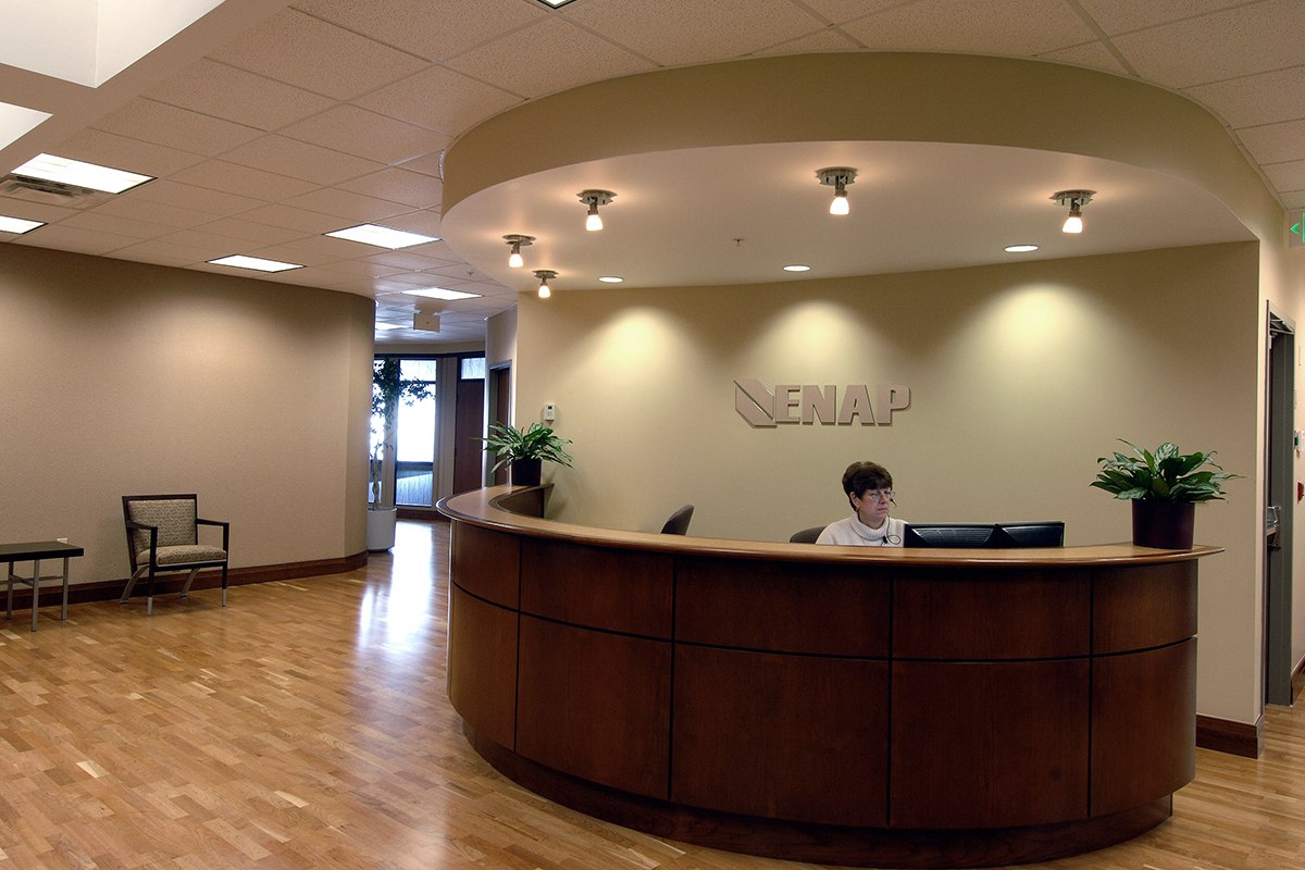 A person sits behind a curved wooden reception desk in a spacious lobby with hardwood floors, light walls, ceiling lights, and potted plants. The word ENAP is mounted on the wall behind the desk.