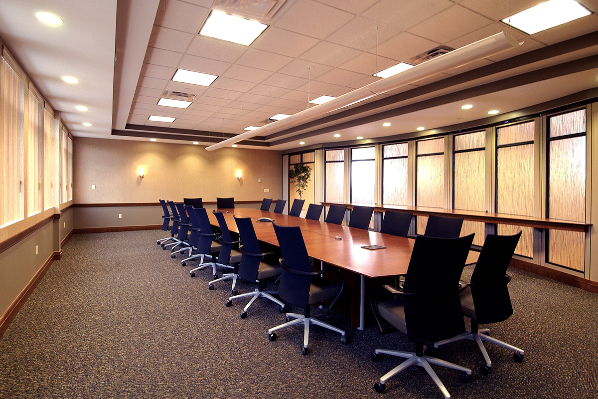 A large, empty conference room with a long wooden table, black swivel chairs, carpeted floor, and windows along one wall allowing natural light.