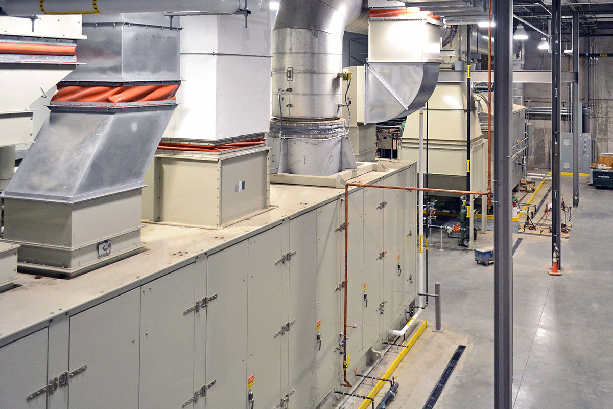 Large industrial HVAC system with metal ducts, ventilation shafts, and control panels in a spacious, well-lit facility with concrete floors and various pipes and equipment visible.
