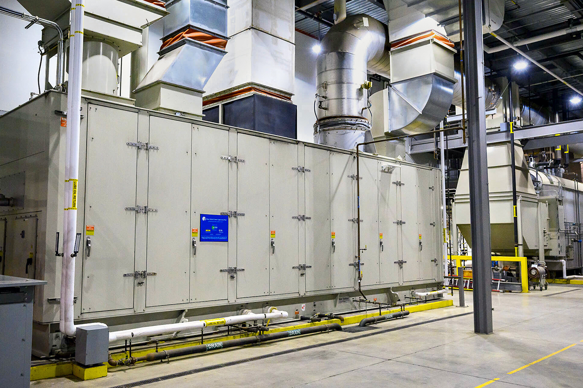 Large industrial air handling unit with multiple doors, pipes, and ducts inside a factory setting. Overhead metal ductwork connects to the machine, and safety barriers line the floor around the equipment.