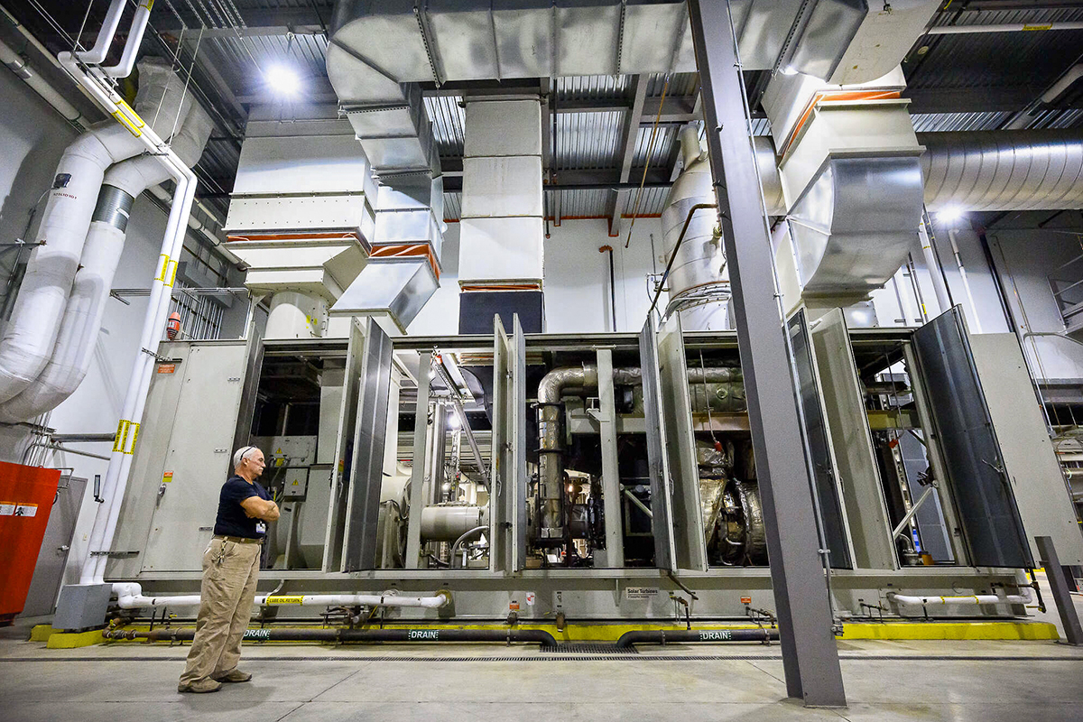 A man stands in an industrial facility, observing large machinery and complex ventilation ducts under bright lighting.
