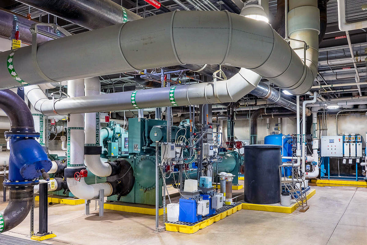 Large industrial room with exposed pipes, machinery, and control panels. Metal ductwork runs across the ceiling, surrounded by equipment such as pumps, tanks, and gauges. The floor is clean and the area is brightly lit.