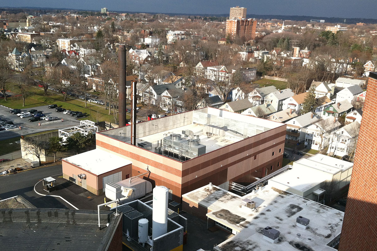 Aerial view of a flat-roofed industrial building with HVAC units, surrounded by residential houses and trees under a cloudy sky.