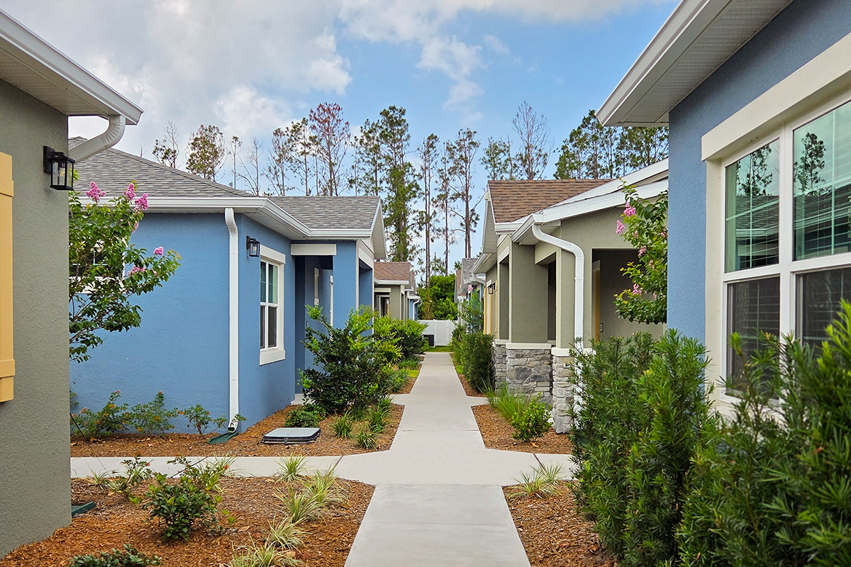 A sidewalk runs between single-story, colorful houses with landscaped gardens and shrubs under a partly cloudy sky.