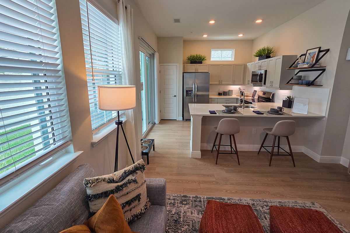 Modern kitchen with white cabinets, island seating for two, stainless steel appliances, and decorative plants. Large windows and a lamp brighten the cozy, neatly arranged living area in the foreground.