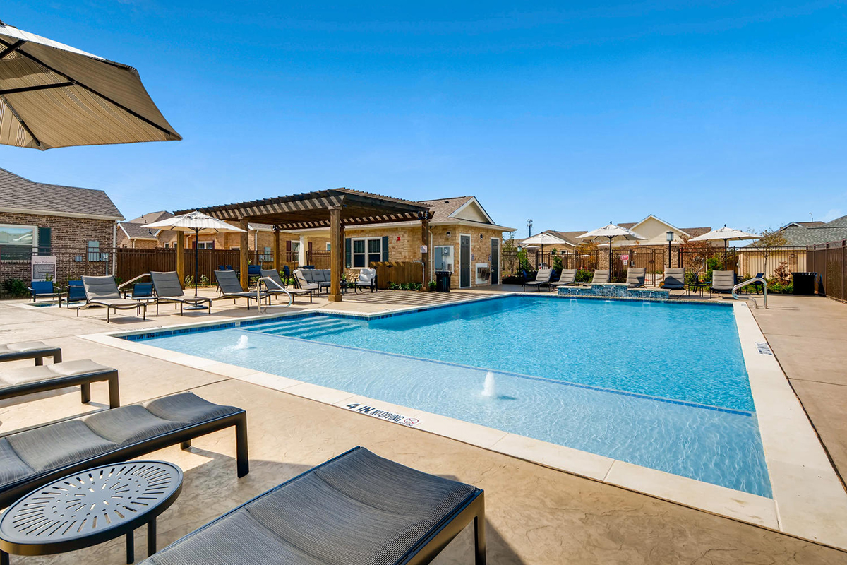Outdoor swimming pool with lounge chairs, pergola, and sun umbrellas on a sunny day. Single-story houses and blue sky are visible in the background. Pool has fountains and steps for entry.