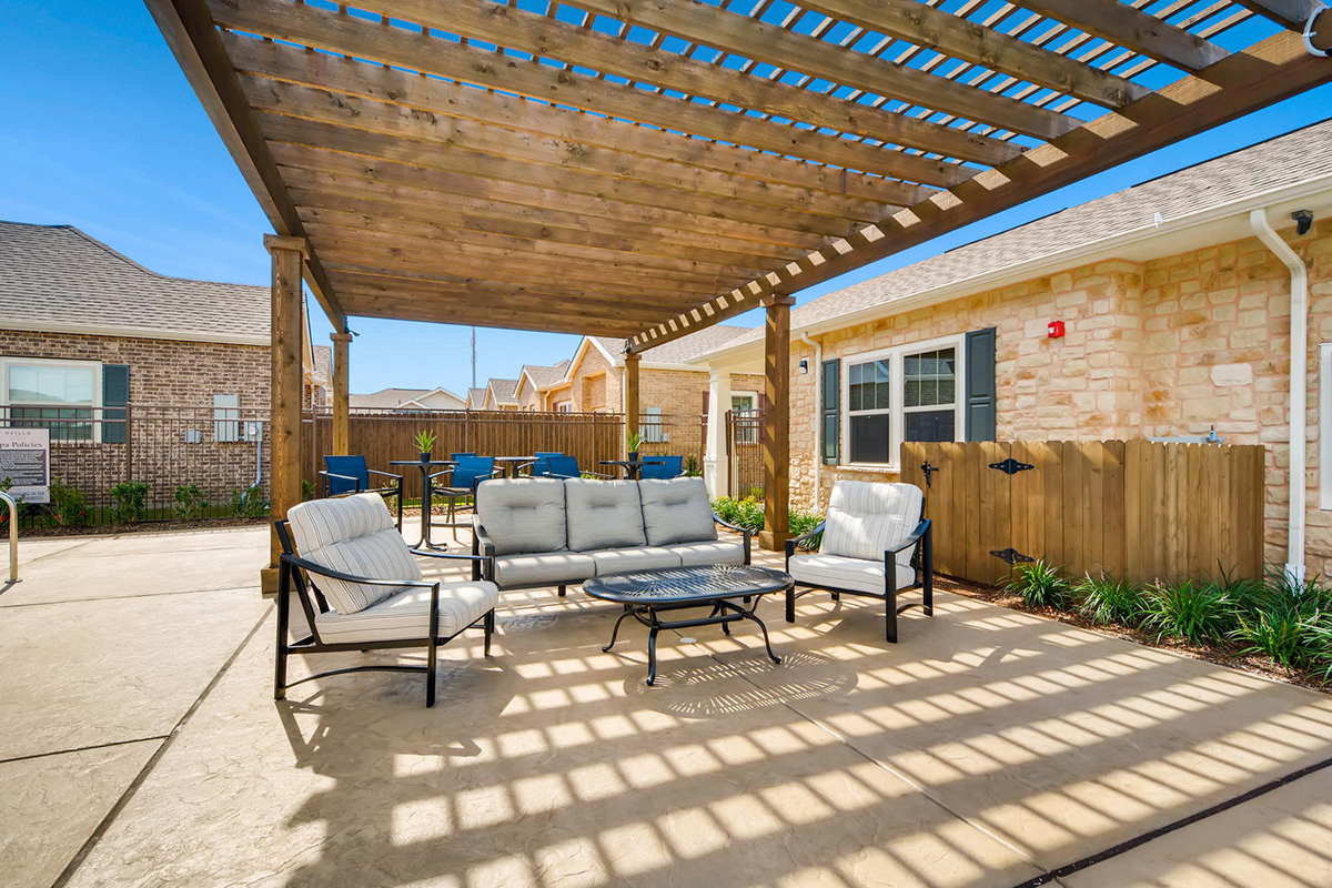 Outdoor patio area with cushioned chairs, a sofa, and a coffee table under a wooden pergola, casting striped shadows on the concrete. Surrounding are buildings with brick and stone exteriors.