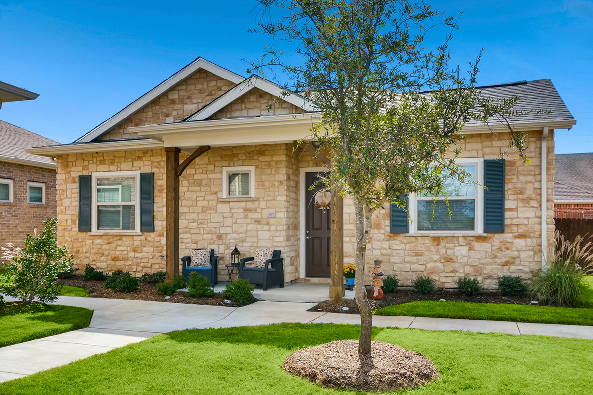 Single-story stone house with blue shutters, a small front porch with chairs, a dark front door, and a landscaped yard with a tree and green grass under a clear blue sky.