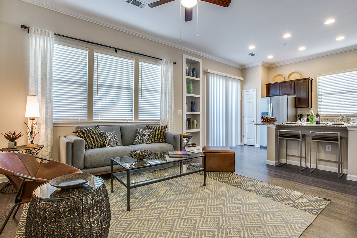 Modern living room with a gray sofa, glass coffee table, patterned rug, two brown chairs, and a kitchen area with barstools. Large windows and sliding glass doors provide natural light.