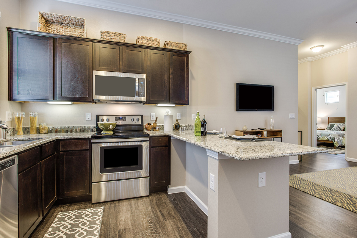Modern kitchen with dark wood cabinets, stainless steel appliances, granite countertops, and a breakfast bar. The living area and bedroom are visible in the background. Baskets are displayed on top of the cabinets.
