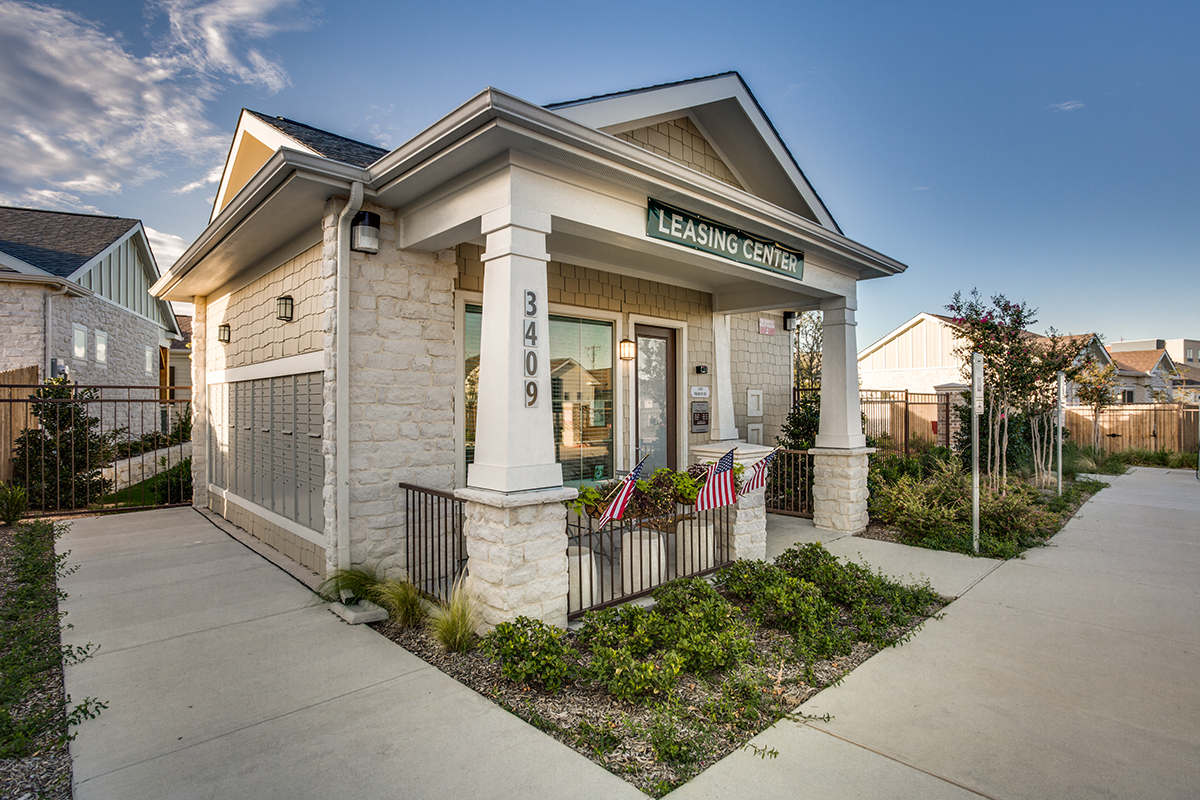 Small stone building labeled Leasing Center with American flags near the entrance, surrounded by sidewalks, greenery, and neighboring houses in the background on a clear day.