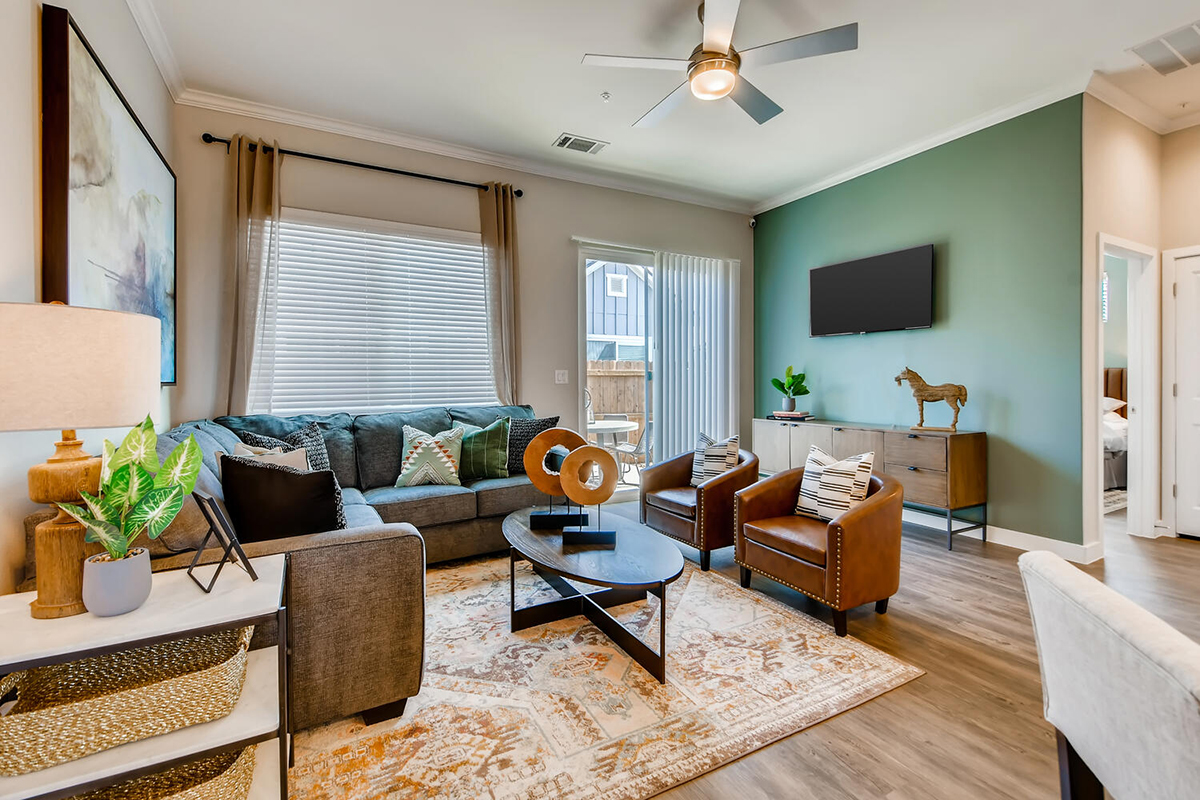 Modern living room with a gray sectional sofa, two brown leather chairs, a coffee table, TV on a green accent wall, and large windows letting in natural light. Decorative items and plants enhance the cozy atmosphere.