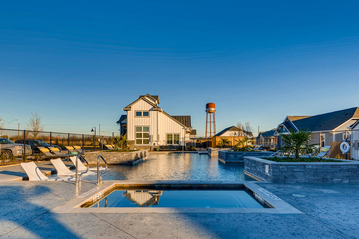 Outdoor swimming pool and hot tub with lounge chairs and a modern farmhouse building in the background on a clear, sunny day. A water tower and additional houses are visible in the distance.