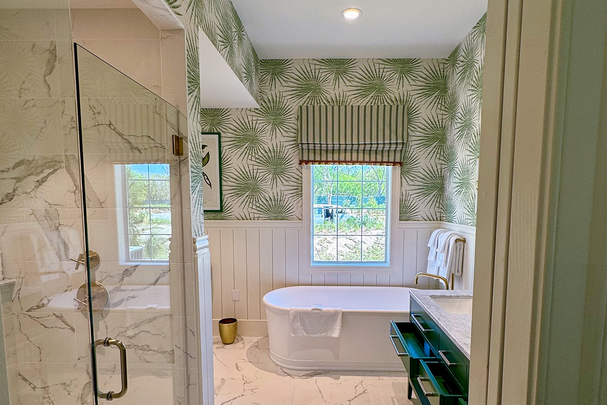 Bright bathroom with palm leaf wallpaper, a freestanding white tub, marble floors, glass shower, window with striped Roman shade, white towels, and gold accents.