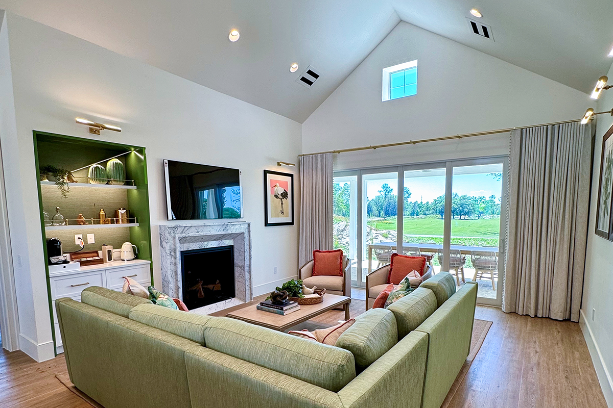 A bright living room with a green sectional sofa, fireplace, wall-mounted TV, two red chairs, and large glass doors opening to a patio with a scenic outdoor view.