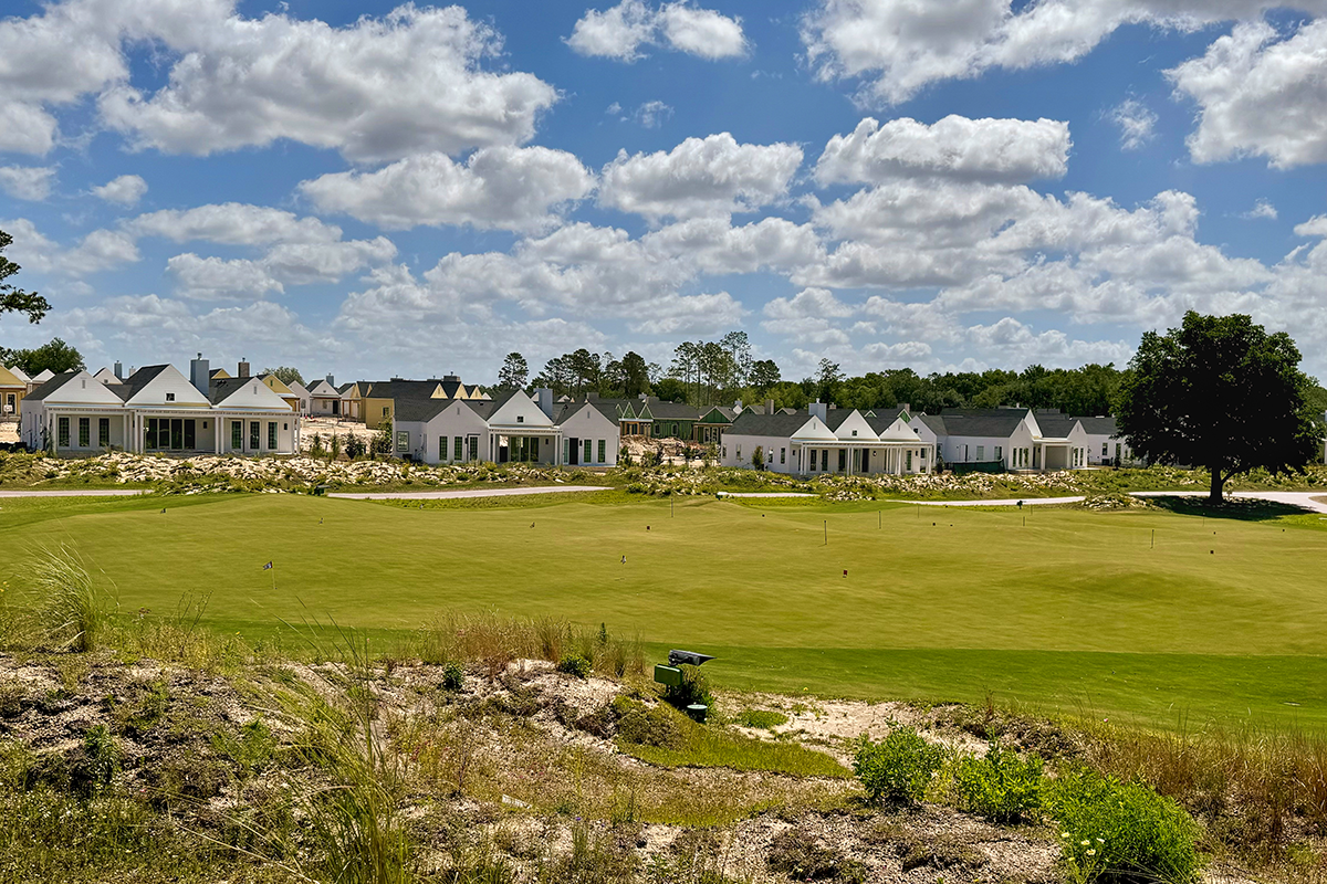 A row of white houses faces a green golf course under a blue sky with scattered clouds, with trees and shrubs in the background.
