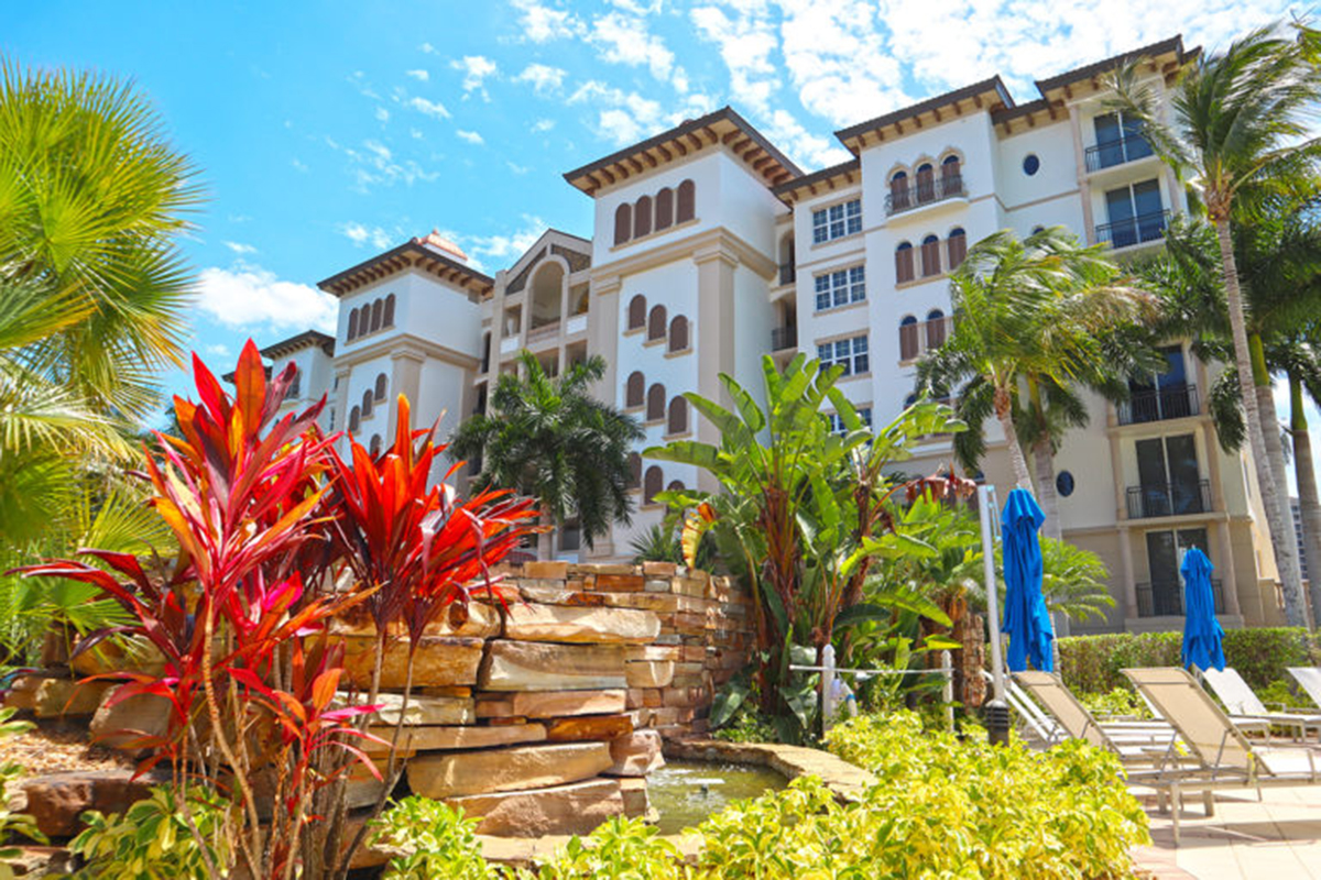 A resort-style building with palm trees, tropical plants, lounge chairs, blue umbrellas, and a decorative stone waterfall under a sunny sky.