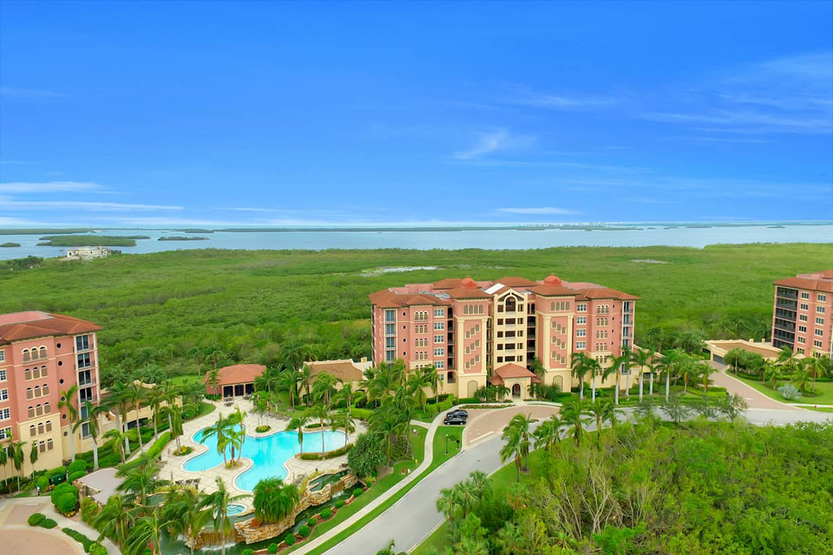 Aerial view of luxury resort buildings surrounded by palm trees, a swimming pool, and lush greenery with water and blue sky in the background.
