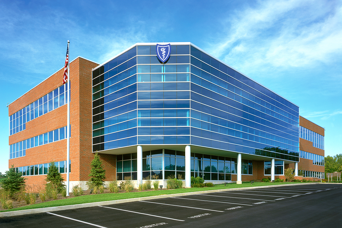 A modern, three-story office building with blue glass windows and a red-brick exterior, an American flag, and an empty parking lot in front under a blue sky.