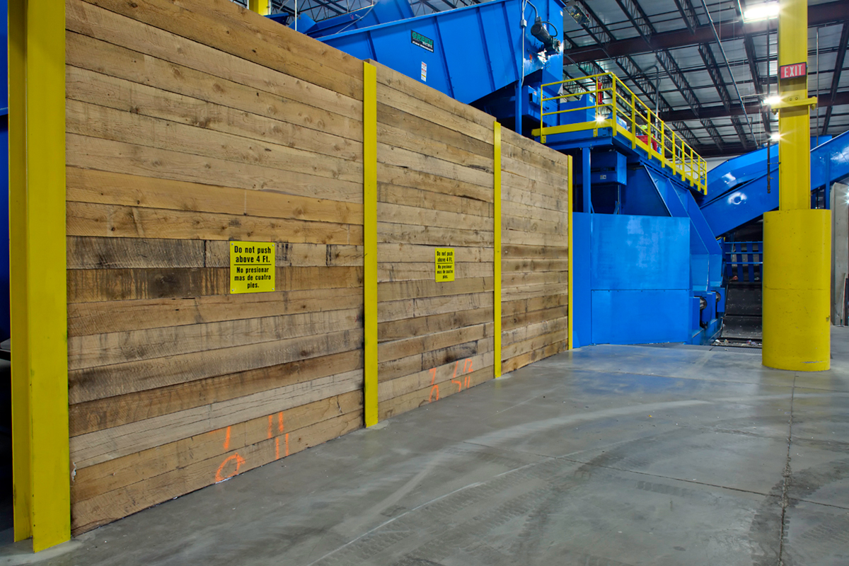 A wooden barrier with yellow warning signs stands inside an industrial facility, near large blue machinery and yellow posts. The floor is concrete, and overhead lights illuminate the space.