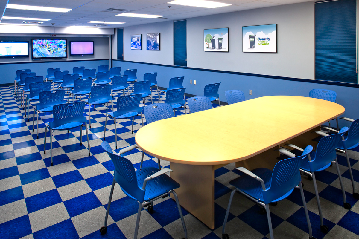A modern conference room with blue chairs, a long wooden table, multiple screens on the wall, checkered blue floor tiles, and framed posters on the walls. The room is well-lit and organized for meetings or presentations.