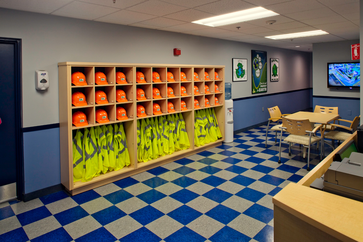 Wall shelves with orange hard hats and yellow reflective safety vests, a table with chairs, safety posters, a TV monitor, and a blue checkerboard floor in a brightly lit room.