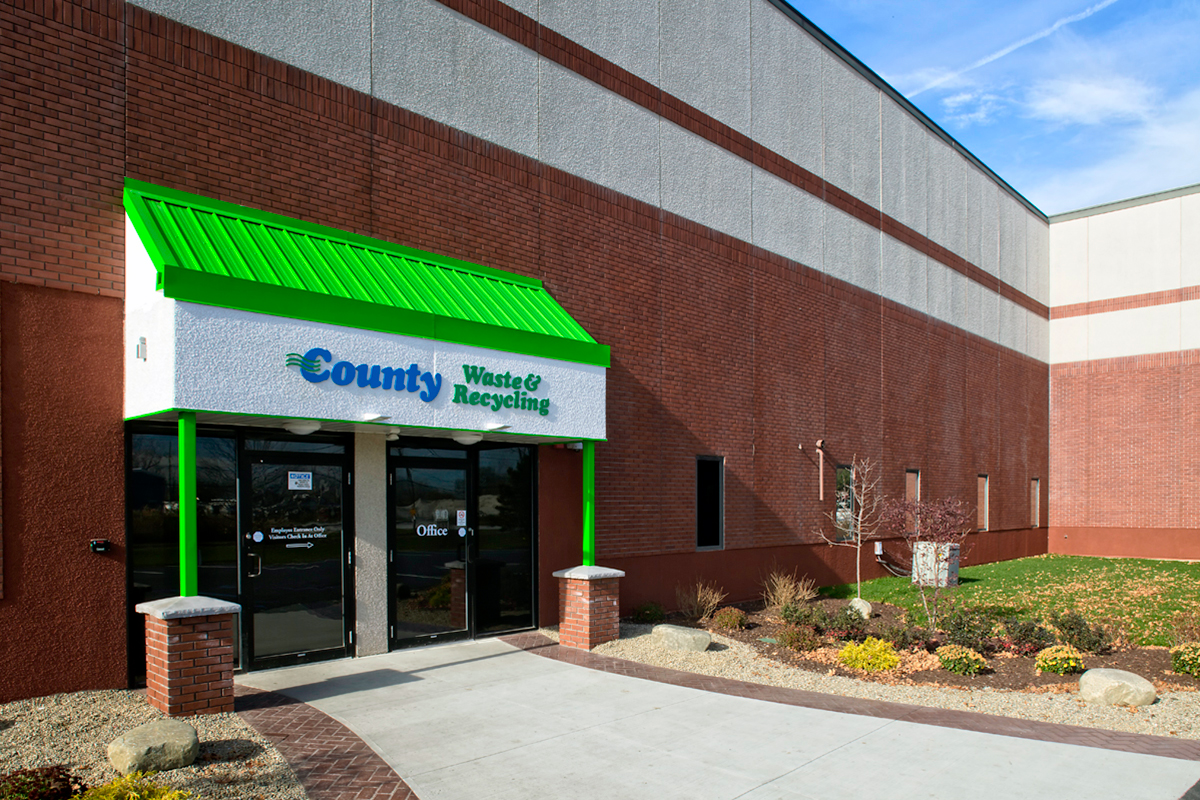 Entrance to a County Waste & Recycling building with glass doors, a green awning, and landscaped grounds featuring rocks, shrubs, and small plants along the walkway.