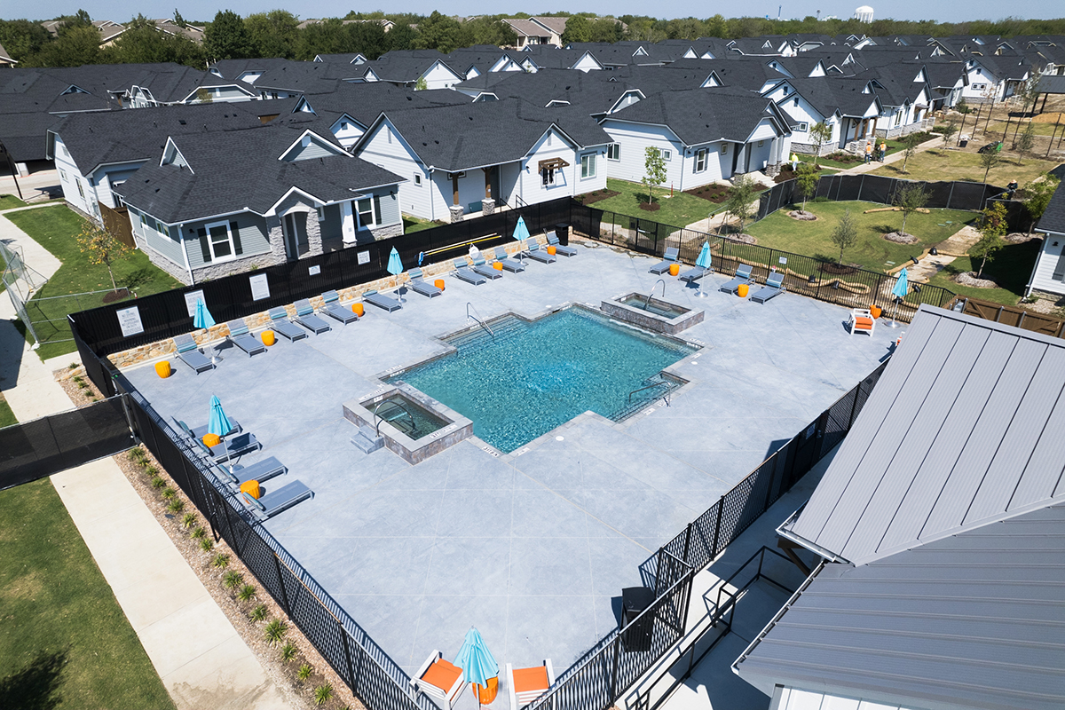 Aerial view of a fenced community pool area with lounge chairs, umbrellas, and a hot tub, surrounded by rows of modern suburban houses.