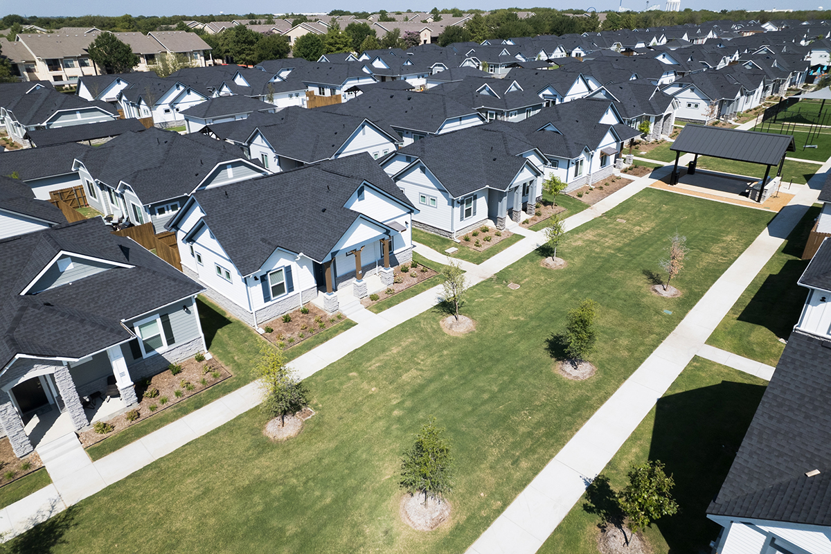 Aerial view of a suburban neighborhood with rows of similar single-story houses, green lawns, young trees, and walkways connecting the homes, under a clear sky.