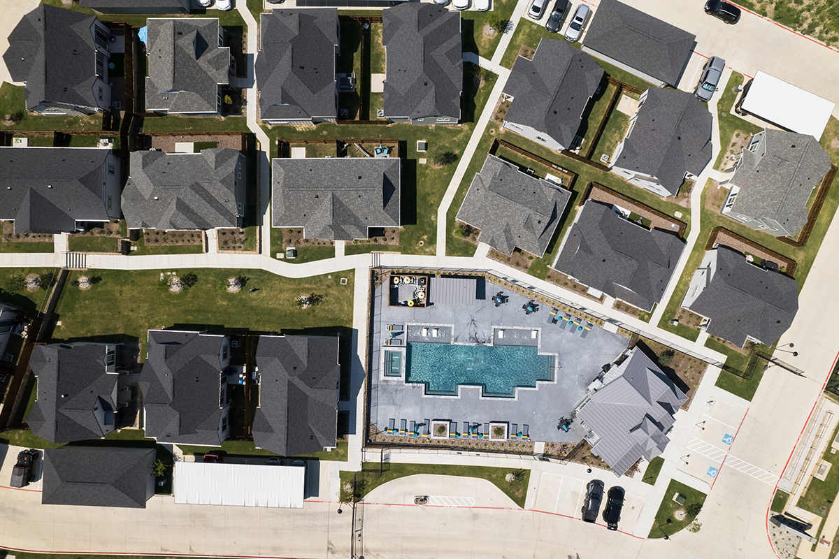Aerial view of a neighborhood with multiple houses, driveways, parked cars, green lawns, and a central rectangular swimming pool surrounded by lounge chairs and cabanas.