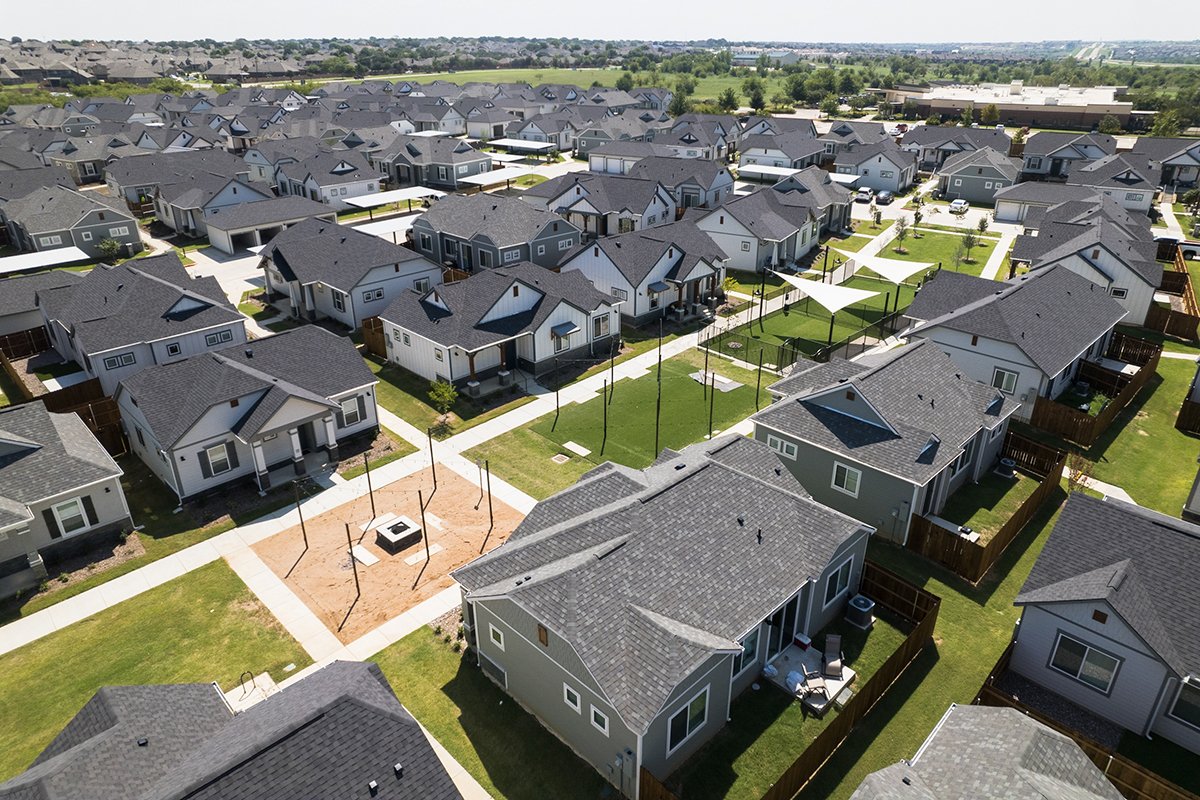 Aerial view of a suburban neighborhood with rows of similar houses, green lawns, sidewalks, backyards, and some fenced areas under a clear sky.