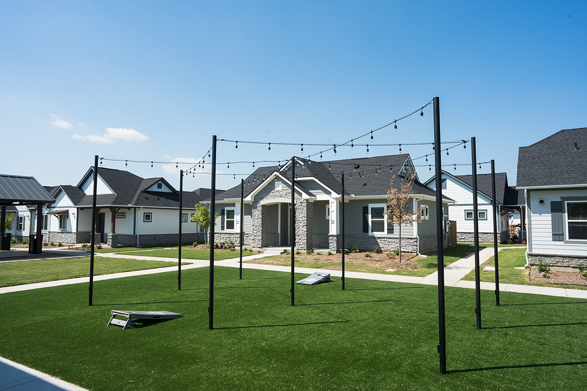 A grassy courtyard with string lights overhead, surrounded by modern single-story houses. Cornhole boards are set up on the lawn under a clear blue sky.