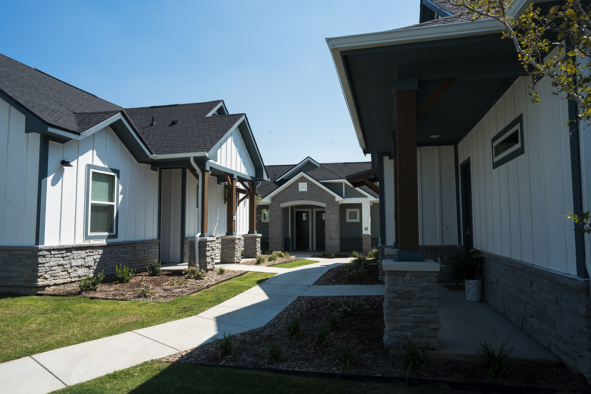Modern single-story houses with white siding, dark roofs, and stone accents are connected by a curved sidewalk and landscaped yards under a clear blue sky.