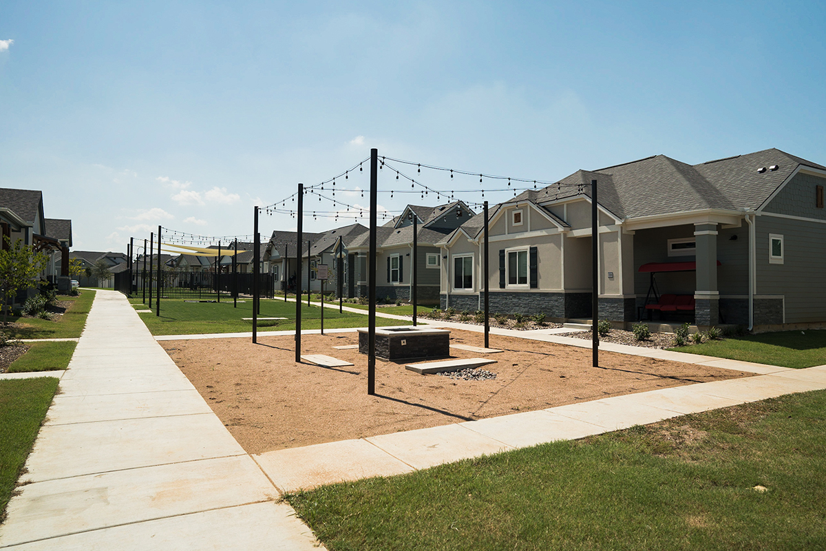 A sandy communal area with string lights, a fire pit, and posts is surrounded by modern single-story houses with well-kept lawns and a wide sidewalk on a sunny day.