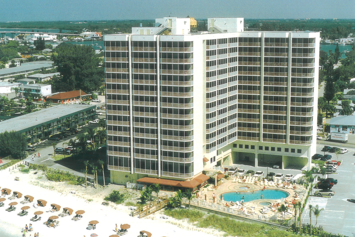 A large beachfront hotel with many balconies faces the ocean. A pool area with lounge chairs and umbrellas sits in front, and the sandy beach is lined with more umbrellas and chairs.