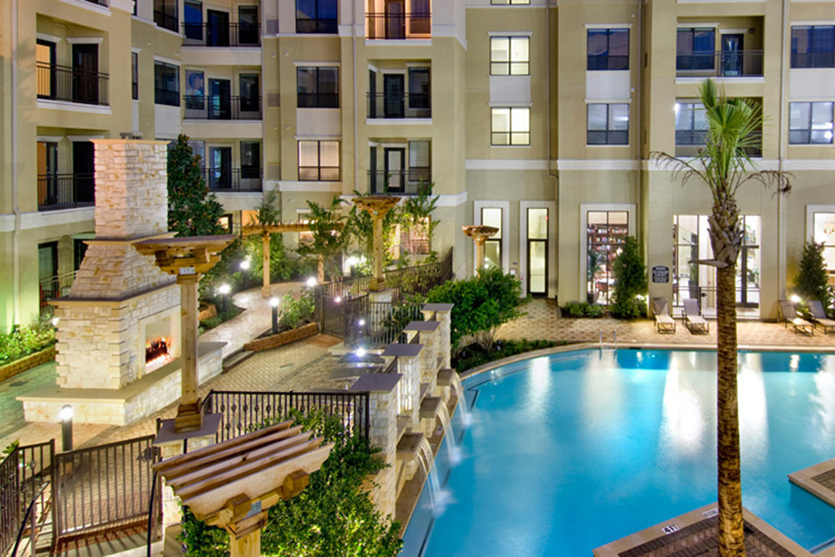 Apartment complex courtyard with a swimming pool, palm tree, outdoor fireplace, pergolas, and landscaped greenery, surrounded by multi-story beige buildings with balconies and large windows at night.