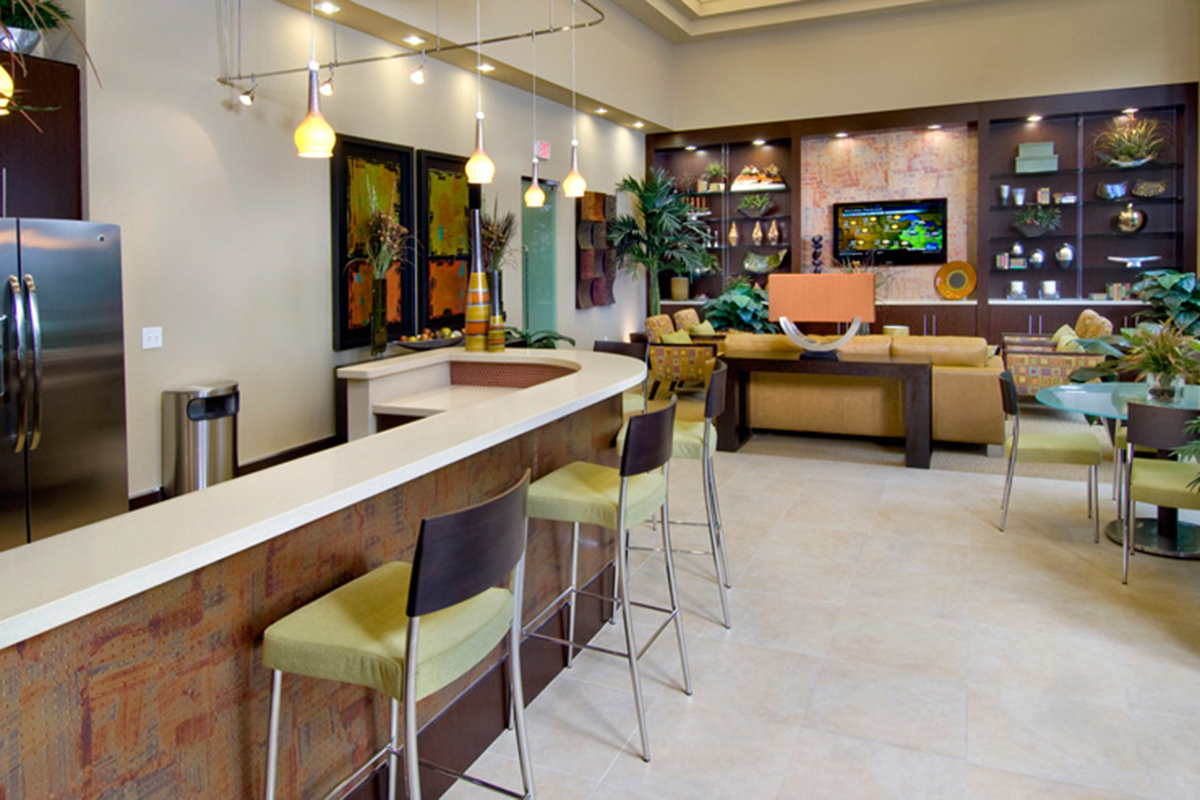 Modern open-concept living space with a kitchen, white countertops, bar stools, a living area with a sofa, TV, bookshelves, indoor plants, and colorful artwork on the walls. Bright lighting gives the room a welcoming feel.