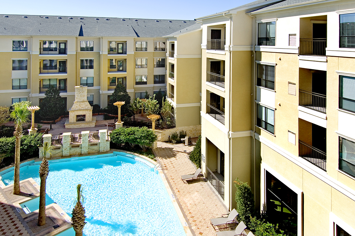Sunny courtyard at an apartment complex with a swimming pool, lounge chairs, palm trees, and a stone outdoor fireplace, surrounded by yellow and cream multi-story buildings.