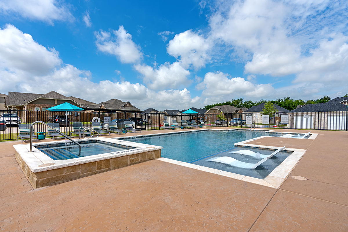 Outdoor swimming pool with lounge chairs partially submerged in shallow water, hot tub on the left, umbrellas and seating area nearby, surrounded by a fence and houses under a partly cloudy sky.