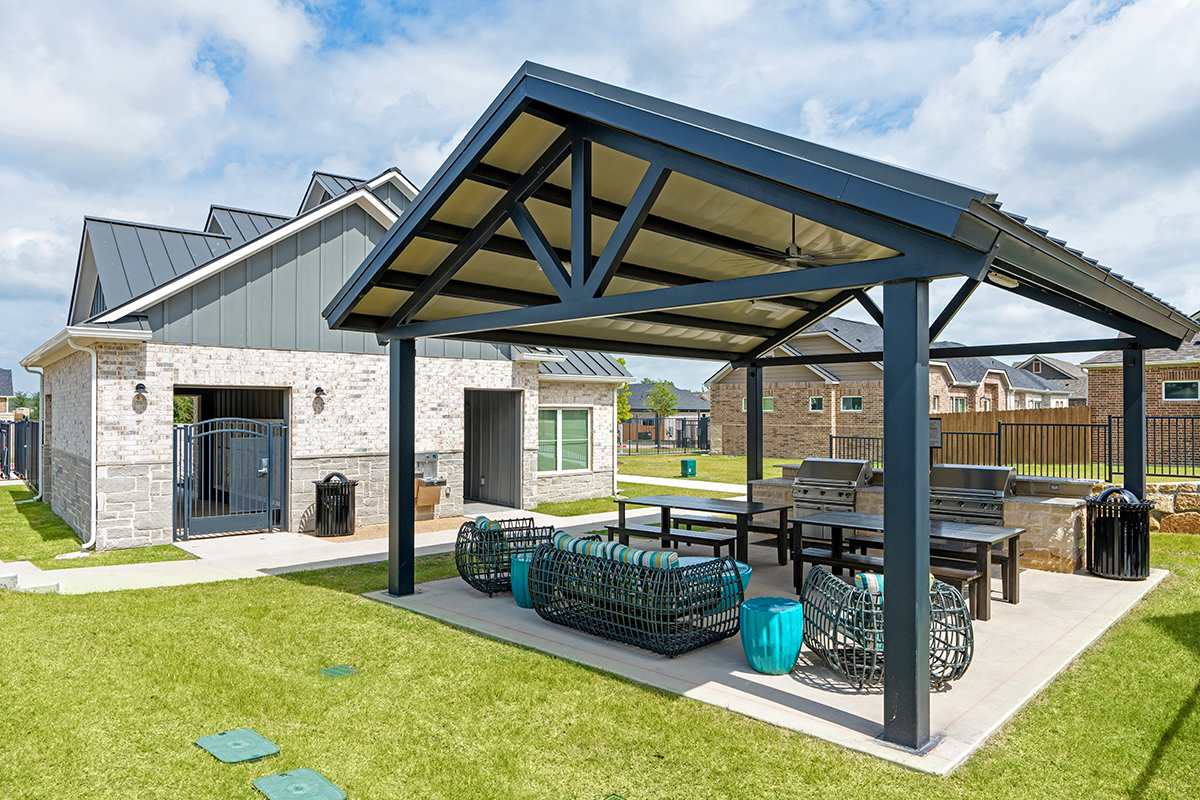 A modern outdoor pavilion with seating, tables, and grills sits on a grassy lawn near a light brick house on a sunny day. Nearby homes and a fence are visible in the background.
