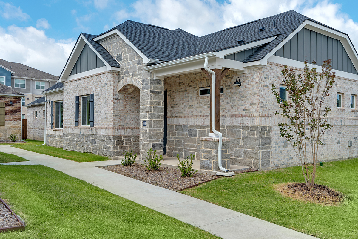 Single-story brick house with gray stone accents, dark roof, and covered entryway. Small bushes and neatly trimmed lawn border concrete walkways leading to the front door.