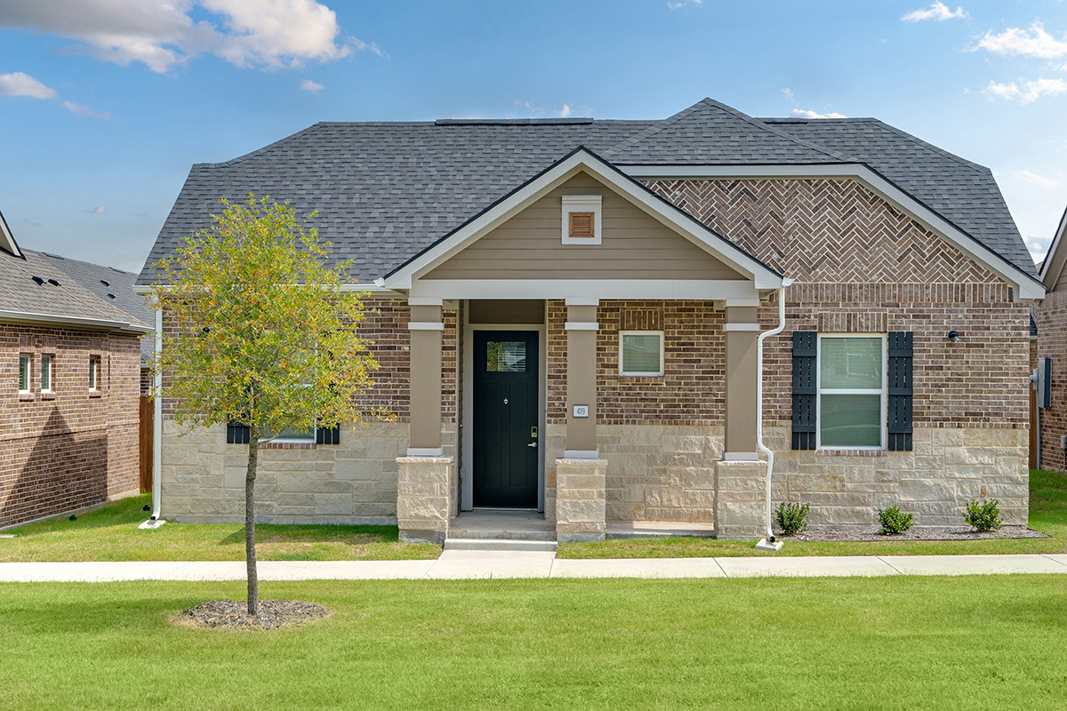 Single-story brick house with a small tree in front, black door, white columns, and neatly trimmed lawn under a blue sky with scattered clouds.