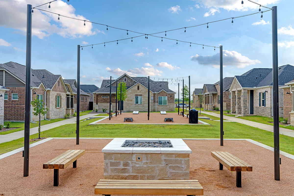 Outdoor communal area with benches around a stone fire pit, string lights overhead, and several brick houses in the background under a blue sky with clouds.
